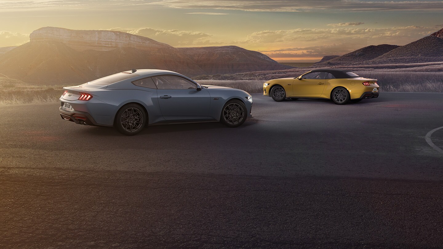 Ford Mustang in Gelb und Grau. Dreiviertelansicht, stehend auf einem Parkplatz bei Sonnenuntergang. Berge im Hintergrund.